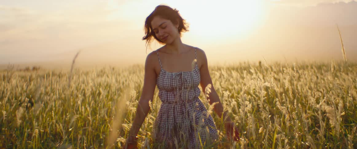 A beautiful young woman in a sundress smiles and walks through a picturesque field of tall grass at golden hour