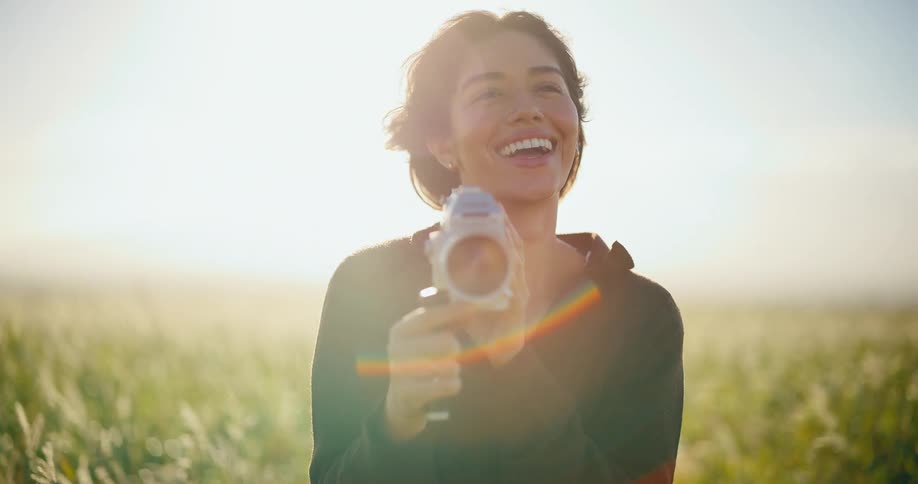 A smiling woman uses a vintage 8mm film camera in a sun-drenched grassy field at golden hour