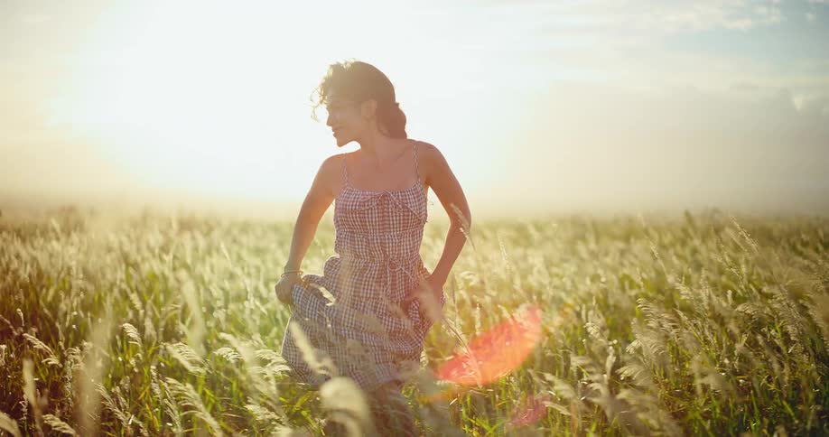 A beautiful woman in a sundress runs joyfully through a sun-drenched field of tall grass in slow motion at sunset
