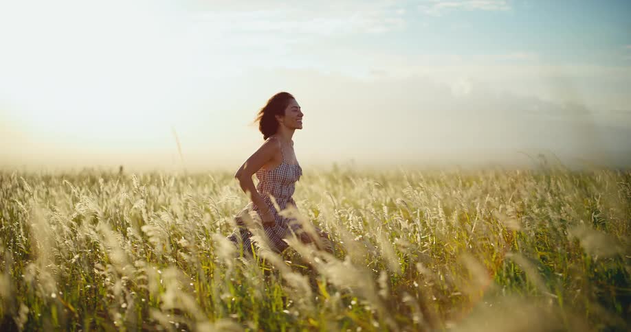 A woman in a flowing dress happily runs through a sunlit field of tall grass as a friend films her with a vintage camera in slow motion