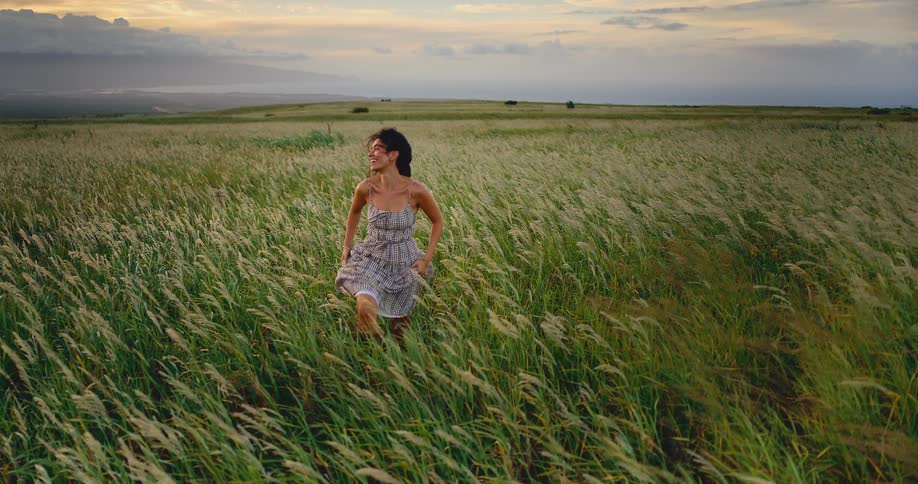 A beautiful young woman runs smiling through a windy field of tall grass at sunset in slow motion