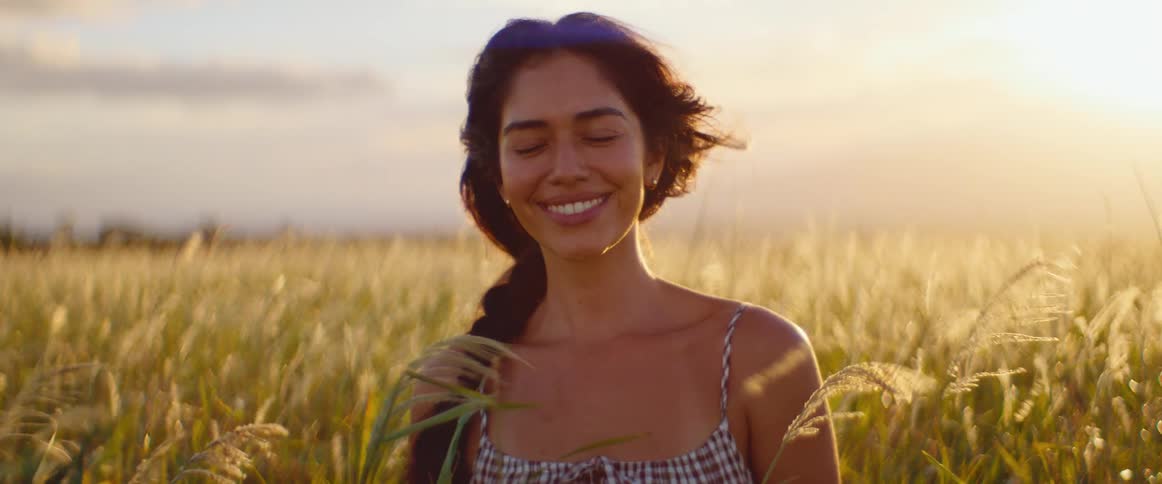 A beautiful young woman smiles and enjoys the warm golden sunlight in a field of tall grass in slow motion