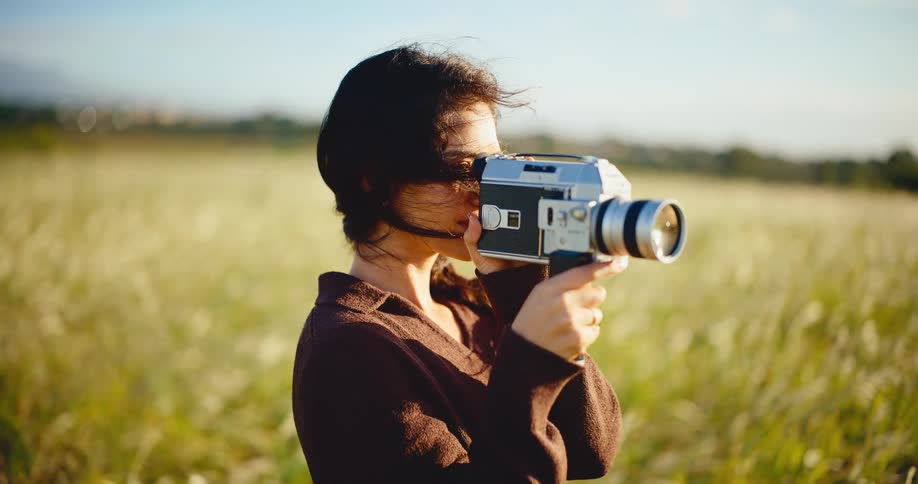 A young woman with windblown hair uses a vintage Super 8 camera to film in a sunny, green field in slow motion