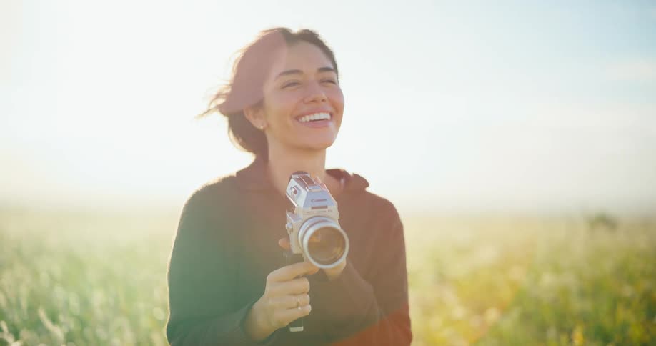 A smiling female filmmaker captures the moment on her vintage 8mm camera in a sunlit field at golden hour