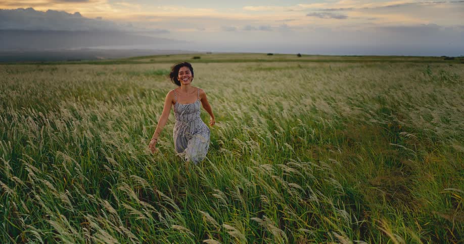 A beautiful young woman runs smiling through a windy field of tall green grass at sunset in slow motion