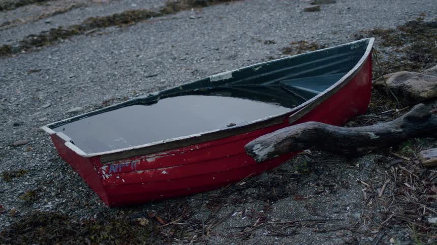 Rain gently falls, creating ripples in the water filling a weathered red rowboat beached on a gravel shore