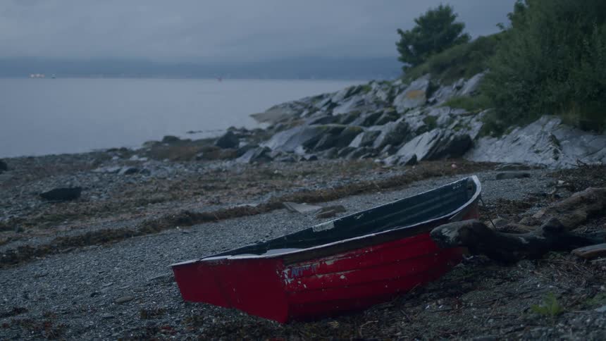 A weathered red rowboat sits on a rocky pebble beach on a quiet, overcast day by the water