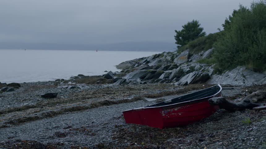An old red boat rests on a desolate rocky beach by the calm sea on a gloomy, overcast day
