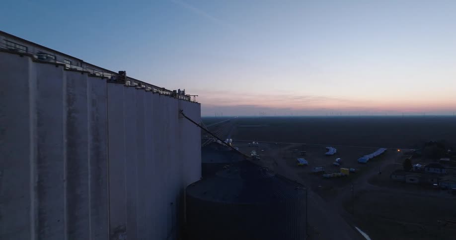 A slow aerial push-in shot captures a massive grain elevator at twilight, revealing the vast rural landscape and a distant wind farm