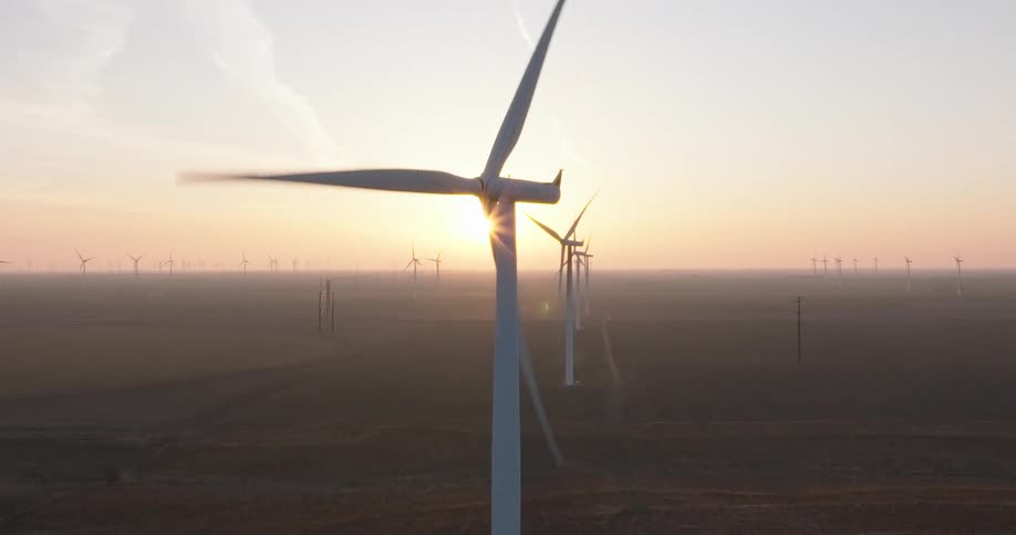 An aerial view of a wind farm shows rows of spinning turbines silhouetted against a beautiful golden sunrise