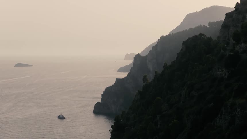 Yachts and sailboats cruise along the dramatic, tree-covered cliffs of the Amalfi Coast during a serene and hazy evening