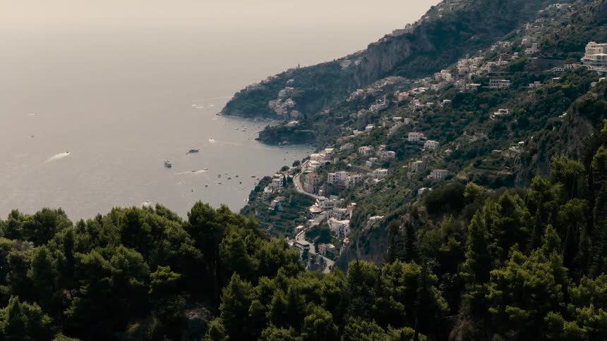 An aerial view pushes over a green forested cliff to reveal the breathtaking coastal town of Positano, Italy, nestled on the mountainside