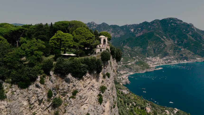 An epic aerial view pulls back from the scenic Terrace of Infinity at Villa Cimbrone, revealing the majestic cliffs and blue waters of the Amalfi Coast
