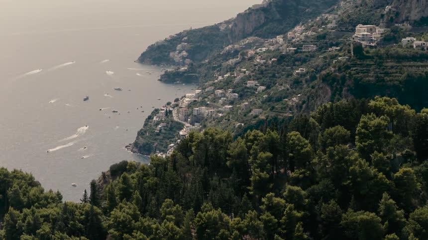 A stunning aerial reveal over a lush green forest unveils the picturesque Amalfi Coast with boats dotting the beautiful blue sea
