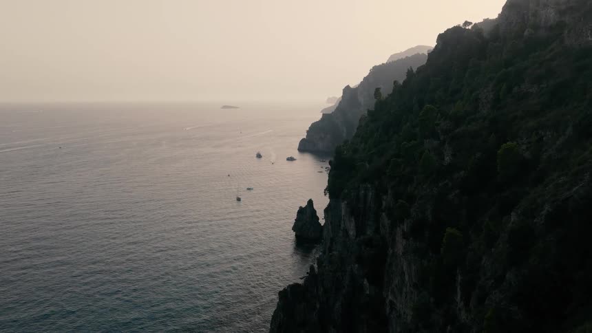 An aerial view slowly reveals the dramatic tree-covered cliffs of a rugged coastline dotted with boats at dusk