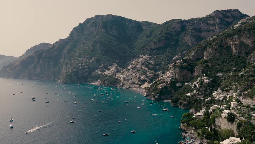 A stunning wide shot of the iconic Amalfi Coast shows the beautiful village of Positano nestled into the cliffs above a bay filled with luxury boats and yachts