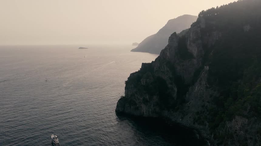An aerial view of yachts and boats near the dramatic, tree-covered cliffs of a rugged coastline during a hazy sunset