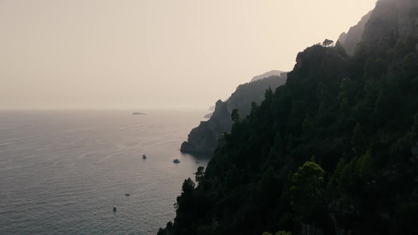 Aerial footage slowly moves along a dramatic, tree-covered cliffside overlooking the ocean and boats at hazy sunset