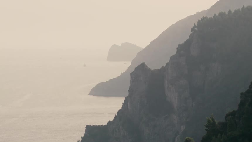 Misty mountains covered in lush green forests rise out of the calm sea along a dramatic, hazy coastline