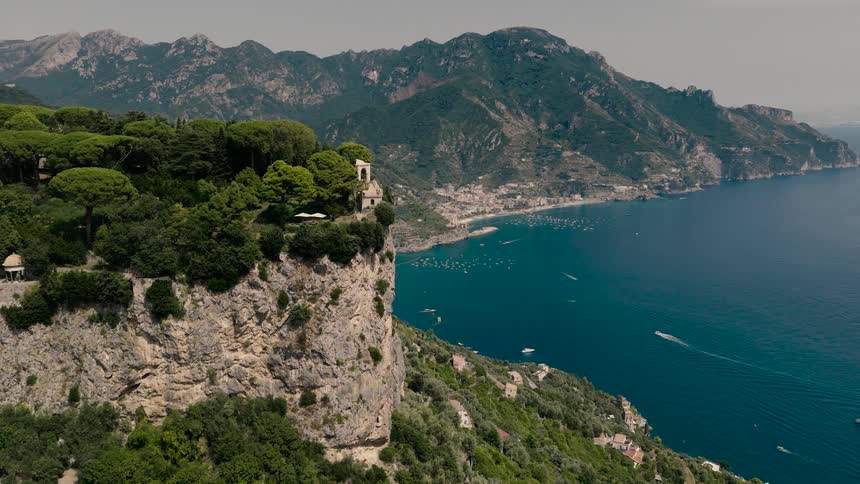 A breathtaking aerial view captures the dramatic cliffs of the Amalfi Coast, featuring a historic villa overlooking the serene blue sea with boats below