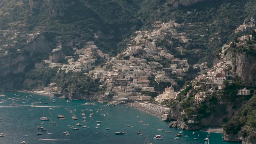 A slow zoom-out shot reveals the stunning coastal town of Positano, Italy, with its colorful houses cascading down the hillside to a bustling beach and bay filled with boats
