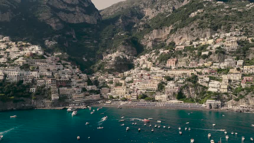 A breathtaking aerial shot pushes towards the iconic cliffside village of Positano, Italy, where boats dot the turquoise bay and a busy beach lines the shore