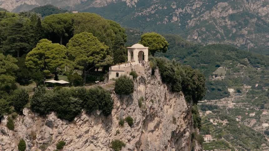 A beautiful view of a structure on a mountain cliff, surrounded by trees and a village on the background