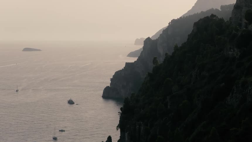 A moody and atmospheric wide shot reveals the rugged, tree-lined cliffs of a coastline with yachts and sailboats on the tranquil sea