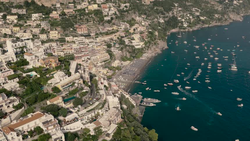 Slow aerial pullback reveals the iconic cliffside town of Positano, with its busy beach and boats dotting the turquoise Amalfi Coast waters