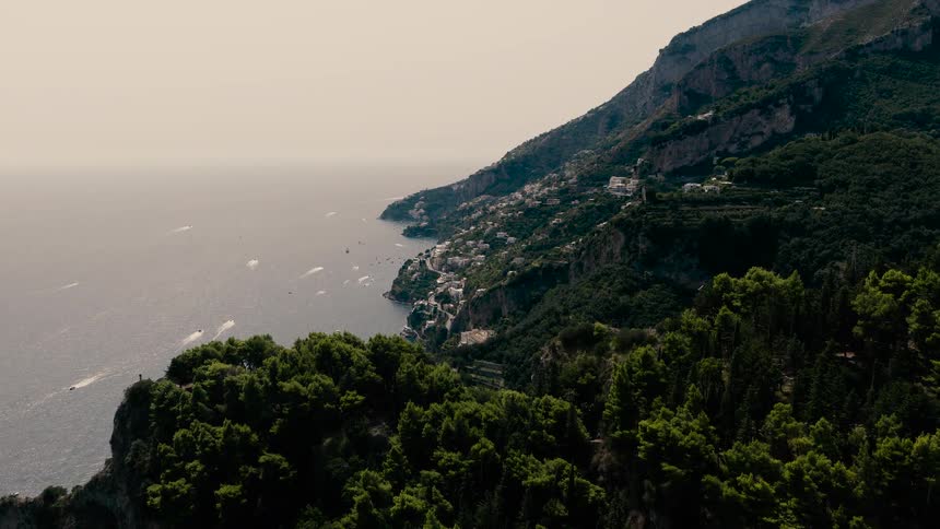 A slow-moving aerial shot flies over green treetops to reveal the beautiful Italian coastline, a bustling marina, and a town built into the mountainside