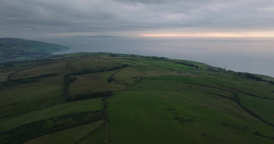 Drone slowly flies over the lush, green rolling hills and patchwork farms along a dramatic coastline at dawn
