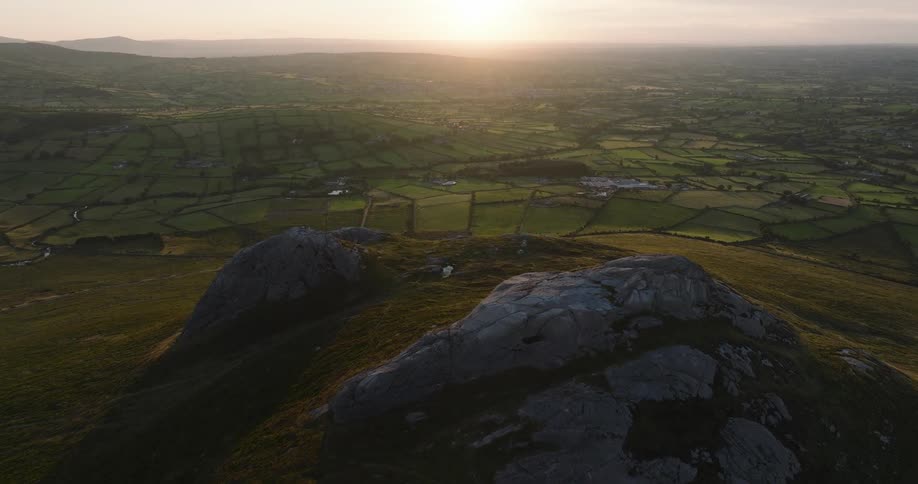 Majestic aerial view of a rugged mountain peak overlooking a sprawling green countryside illuminated by the warm light of a setting sun