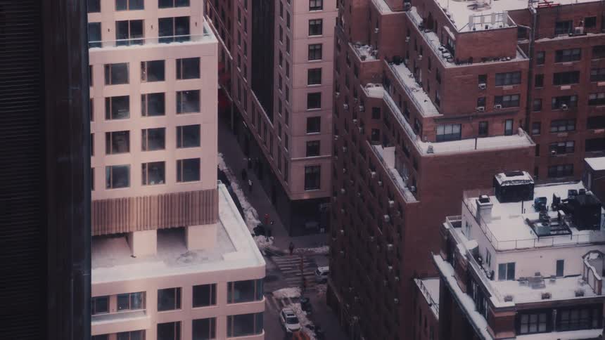 An aerial view captures a quiet, snow-covered city street as steam rises between towering buildings on a cold winter day