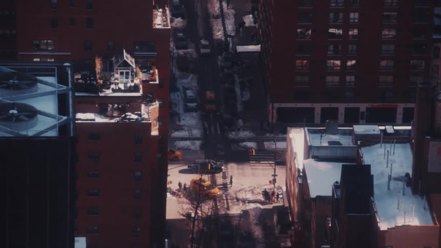 A high-angle view of a snowy New York City street with pedestrians and yellow cabs moving through an intersection at dusk