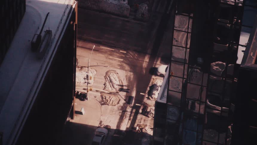 A high-angle aerial view shows traffic moving through a sunlit city street canyon surrounded by snow-covered rooftops