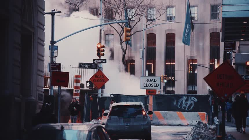 Pedestrians and cars navigate a steamy, closed-off city street with construction signs and snow on a cold winter day
