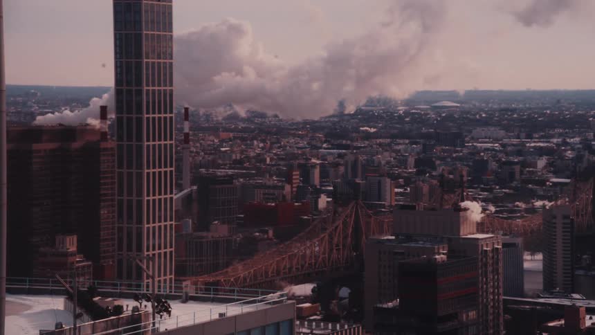 A wide, static shot captures the industrial skyline of New York City, featuring the Queensboro Bridge and billowing smoke from chimneys on a cold day