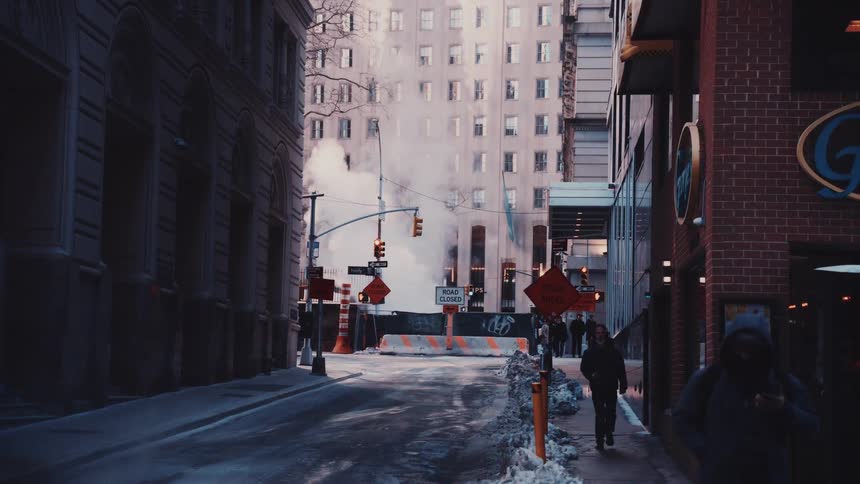 A moody, slow-motion view of a snowy city street in winter, where pedestrians walk past billowing steam from a construction zone