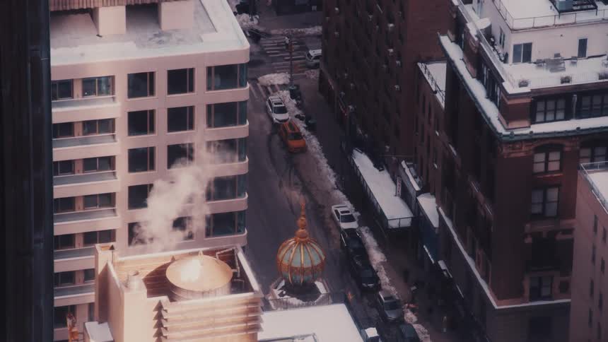 A high-angle view of a snowy New York City street with steam rising from a rooftop as yellow cabs drive below