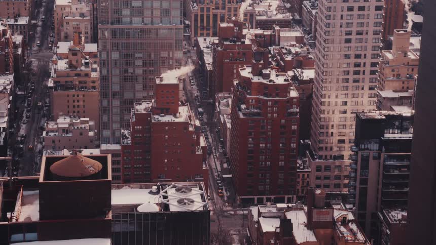 A static high-angle view captures the snow-covered rooftops of New York City in winter as steam rises from a chimney