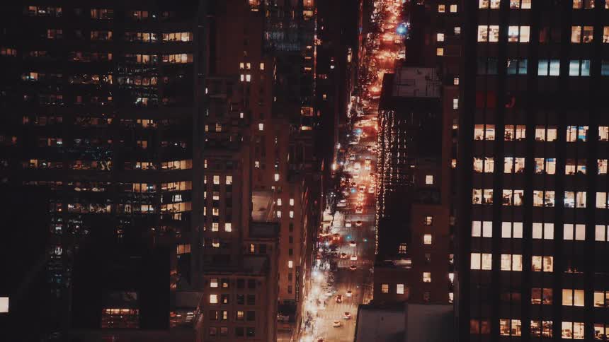 A high-angle static view looks down on a bustling city street at night, where streams of traffic flow between skyscrapers with warmly lit windows
