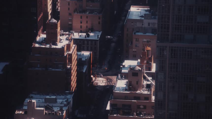 A high-angle view looks down on a city street where sunlight illuminates snow-dusted rooftops while traffic moves in the deep shadows below