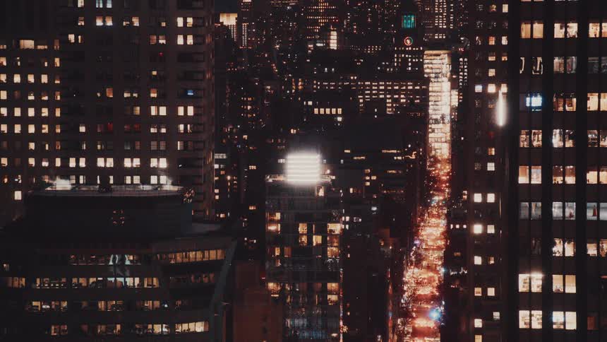 High-angle static shot of a bustling New York City street at night, with traffic flowing between buildings with glowing windows