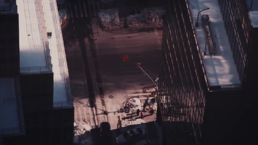 A top-down aerial view captures the flow of traffic and pedestrians on a snowy urban street canyon as long shadows stretch across the pavement