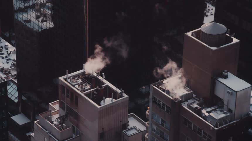 Steam rises from vents on snow-covered city rooftops during a cold winter day