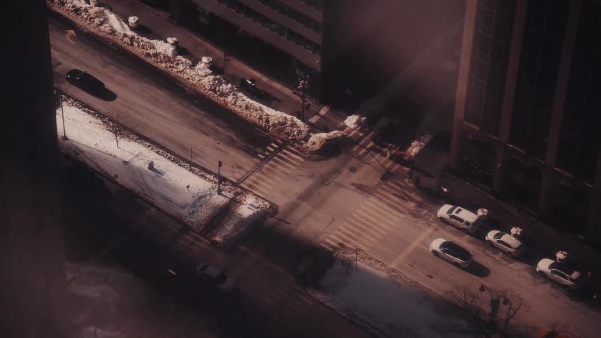 A high-angle time-lapse of a snowy city intersection as cars and taxis drive through casting long shadows in the winter sun