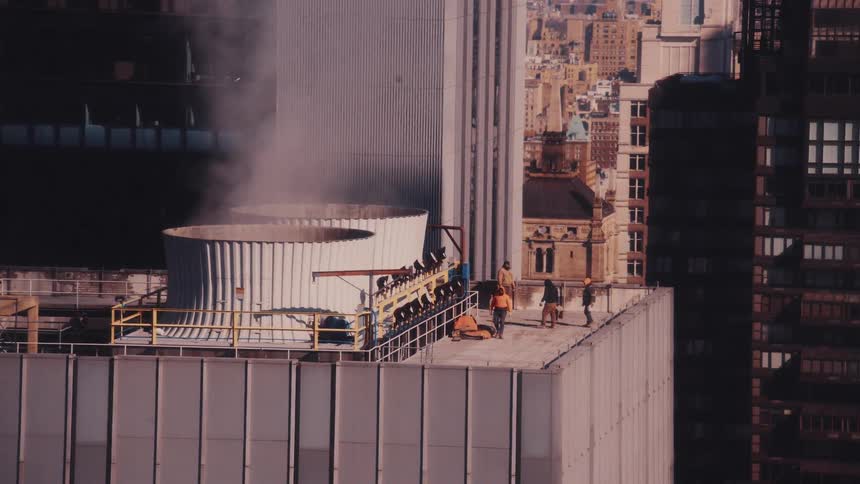 Maintenance workers walk on the rooftop of a skyscraper next to a large steaming ventilation unit with the city in the background