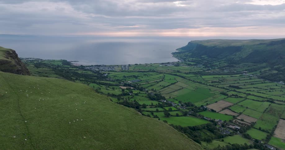 A sweeping aerial drone shot reveals the majestic green hills and coastal valley of Northern Ireland at dawn