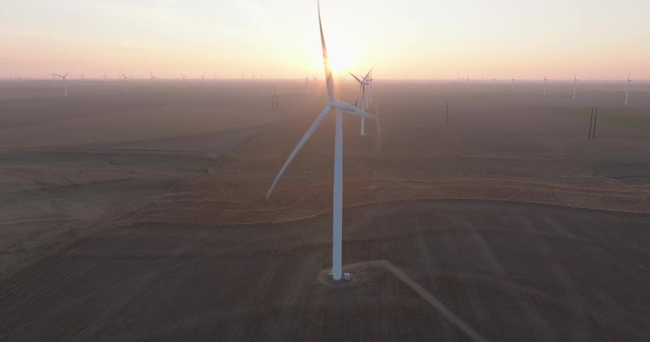 A serene aerial view captures dozens of wind turbines spinning across a vast, hazy landscape during a beautiful sunrise
