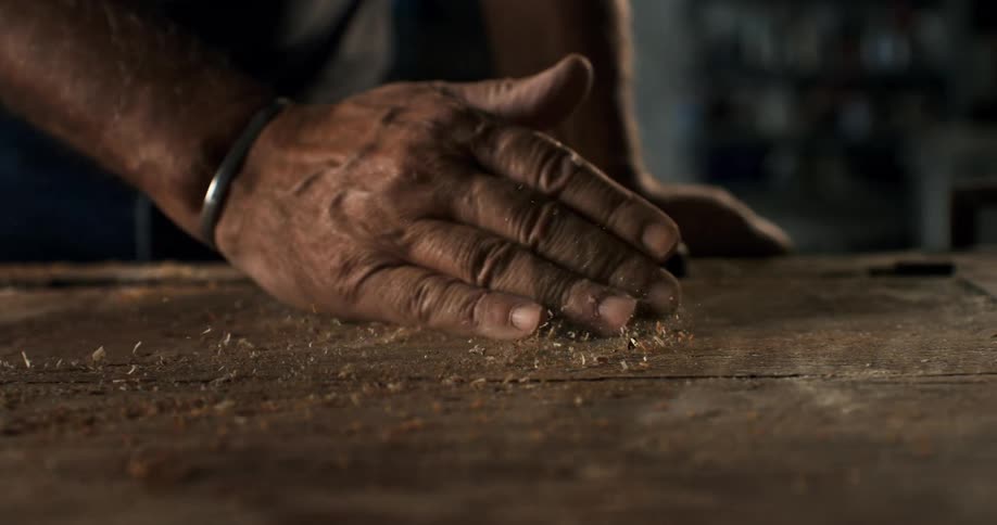 An experienced craftsman's weathered hand slowly sweeps sawdust from a wooden workbench in a workshop